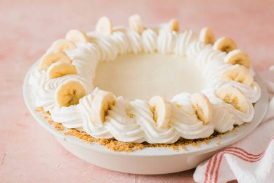 Whole banana cream pie with whipped cream swirls on top and banana slices set against a pink background.