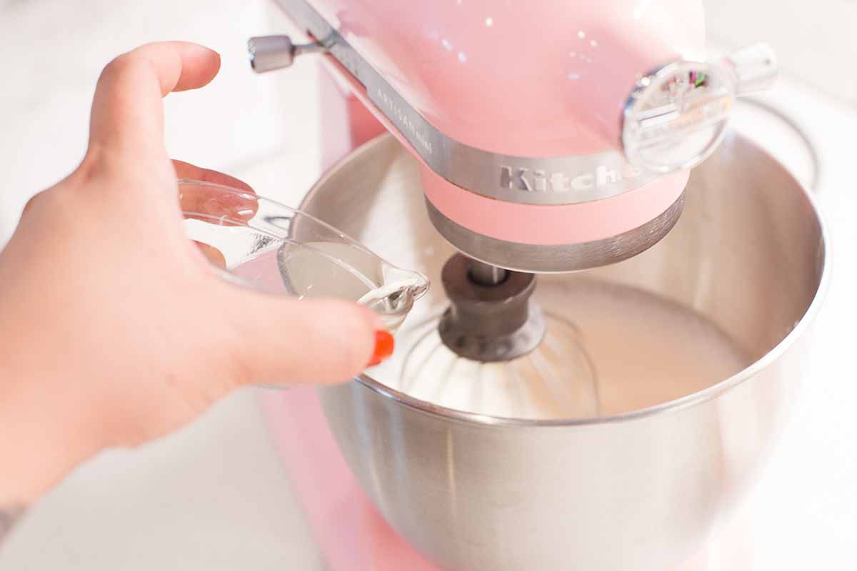 Adding melted gelitin to the bowl of pink standmixer filled with heavy cream.
