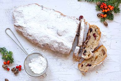 Stollen Christmas Bread filled with rum-soaked dried fruit coated with powdered sugar and sliced on a platter with a fine mesh sieve with powdered sugar in it nearby.