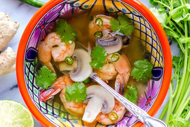 Thai soup with shrimp in a brightly patterned bowl with a silver spoon inside. Sliced lime, ginger, peppers and cilantro stems suround the bowl of light broth, shrimp, mushrooms and cilantro.
