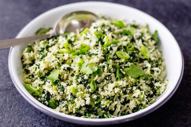 A horizontal image of a bowl of low carb cauliflower rice with sliced scallions and cilantro scattered on the top. A silver serving spoon is in the bowl and the bowl is set on a slate background.