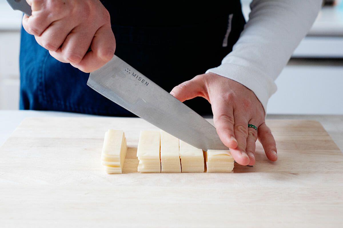 A person with a blue apron and long sleeved white shirt is holding a chef's knife and chopping swiss cheese on a wooden cutting board.