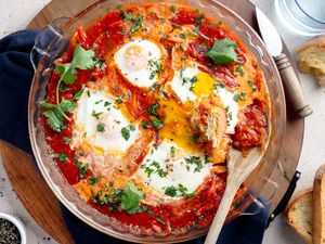 Microwave Shakshuka in a Circular Pyrex Dish on a Circular Tray Surrounded by Slices of Bread on the Counter, a Small Bowl of Cracked Pepper, and a Glass of Water