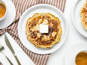 Plate with a Stack of 3-Ingredient Banana Pancakes with Butter, Surrounded by a Plate with More Pancakes, Cups of of Coffee, and Utensils on a Table Napkin