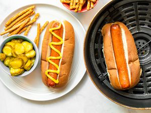 A Plate of Air Fryer Hot Dog in a Bun Topped with Mustard and a Side of Pickles and Pretzels. Next to It Is the Air Fryer Basin with Another Hot Dog in a Bun and a Bowl of Pretzels.