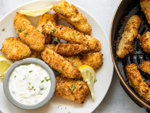 Air Fryer Fish Sticks on a Plate with a Small Bowl of Tartar Sauce and a Lemon Slice. More Fish Sticks in Air Fryer Basin Next to Plate.