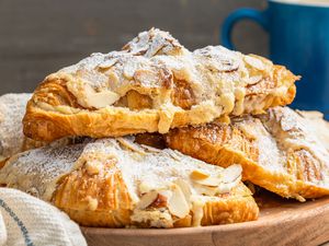Pile of Almond Croissants on a Wooden Tray, and in the Surroundings, a Kitchen Towel and a Blue Coffee Mug