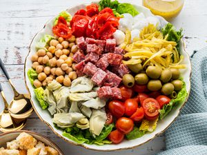 Bowl of Antipasto Salad With the Ingredients in Neat Piles, and in the Surroundings, a Robbin Egg Colored Kitchen Towel, a Bowl of Croutons, a Jar of Dressing, and Two Spoons on the Counter