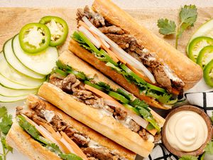 Easy Pork Bánh Mì Surrounded by Slices of Cucumbers and Jalapenos and a Bowl of Mayo