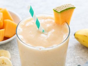 Glassful of Cantaloupe Smoothie With a Slice of Cantaloupe on the Glass Rim, and in the Surroundings, a Bowl of Cantaloupe and Slices of Bananas and a Whole Banana on the Counter