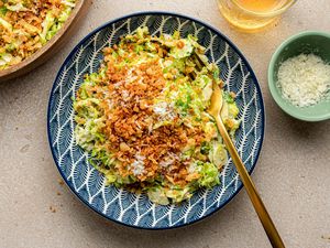 Serving of Brussels Sprouts Caesar Salad with Lemony Breadcrumbs in a Bowl with a Fork Next to a Larger Bowl with More, a Bowl with Shredded Parmesan, and a Glass