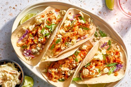 Buffalo Chicken Tacos Topped With Blue Cheese Crumbles and Green Onion Slices and Next to a Lime Quarter, All on a Platter, and in the Surroundings, Another Lime Slice on the the Counter, a Small Bowl With More Cheese, and a Glass of Water