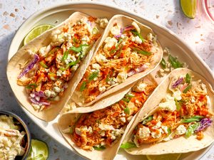 Buffalo Chicken Tacos Topped With Blue Cheese Crumbles and Green Onion Slices and Next to a Lime Quarter, All on a Platter, and in the Surroundings, Another Lime Slice on the the Counter, a Small Bowl With More Cheese, and a Glass of Water