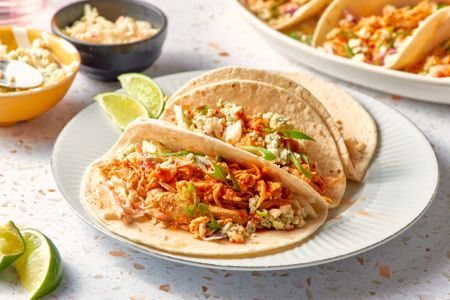 Plate With a Serving of Buffalo Chicken Tacos Topped With Blue Cheese Crumbles and Green Onion Slices and Next to Two Lime Slices. In the Surroundings, a Platter With More Tacos, Lime Slices on the Counter, a Bowl of Coleslaw, and a Glass 