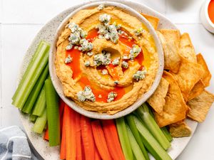 Bowl of Buffalo Hummus on a Plate with Celery Sticks, Carrots Sticks, and Pita Chips, Next to a Small Bowl with Buffalo Sauce 