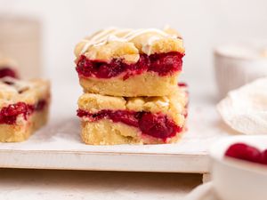 Two Cherry Pie Bars Stacked Together Next to Another Bar, All on a Tray, and in the Surroundings, a Bowl of Cherries and a Bowl of Icing