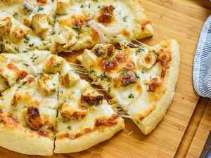 Chicken Alfredo Pizza Cut Into Slices With One Slice Slightly Removed From the Rest to Get a Cheesy Pool, All on a Cutting Board, and Next to the Board, a Blue and White Striped Kitchen Towel