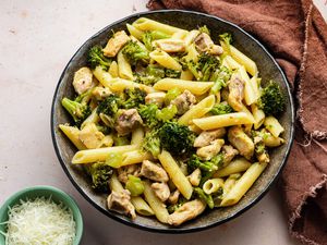 Bowl of Chicken and Broccoli Pasta Next to a Kitchen Towel and a Small Bowl of Cheese