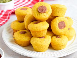 Pile of Corn Dog Muffins Stacked on a Plate, and in the Background, a Red and White Checkered Kitchen Towel and Small Bowls of Toppings (Relish and Ketchup)