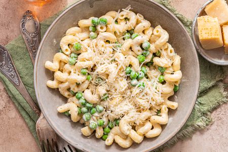 Bowl of Creamy Cavatappi Topped With Shredded Parmesan Next to a Small Bowl With Blocks of Parmesan and Two Forks, All on an Olive Green Kitchen Linen