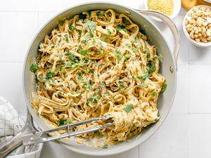 Pan of Super Creamy Vegan Pasta Topped with Parsley and Some Wrapped Around a Pair of Tongs. Next to the Pan, Two Bowls of Ingredients (One: Cashews and Two: Nutritional Yeast)