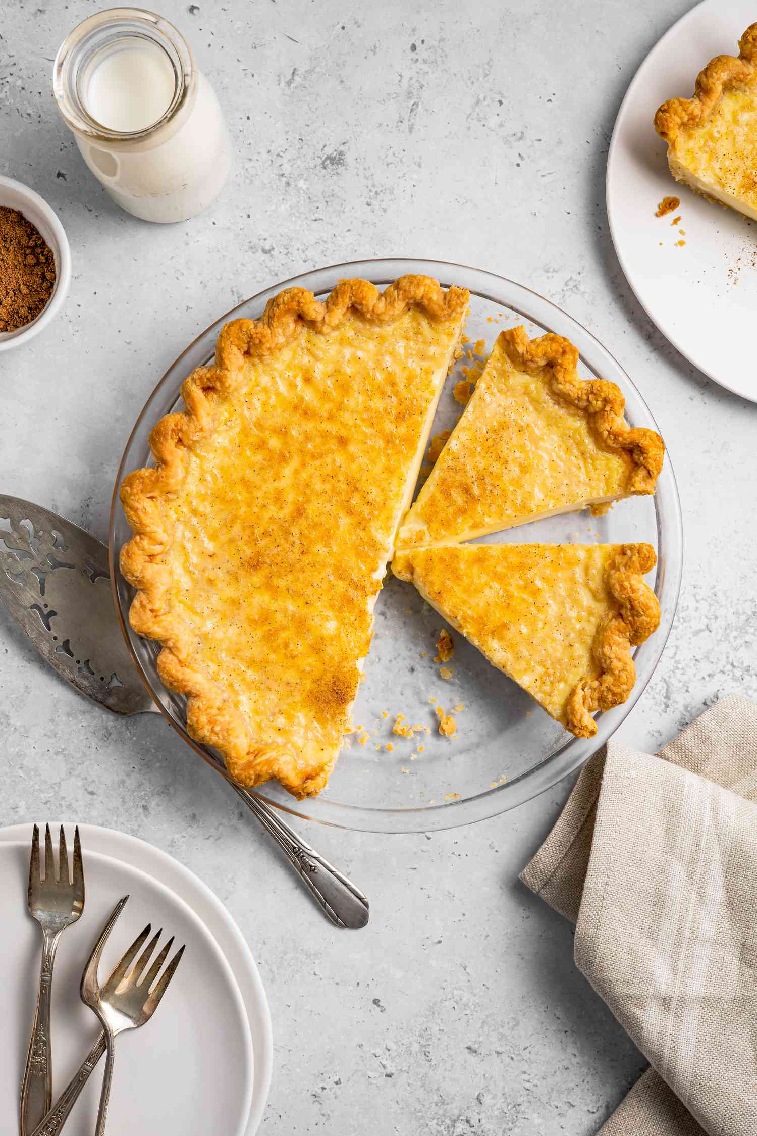 Egg Custard Pie Cut into Slices Next to a Pie Server, Jar of Milk, and Plate with Forks