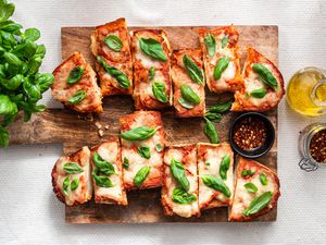 Sliced French Bread Pizza Topped With Fresh Basil (Unsliced) and a Small Bowl of Crushed Peppers on a Wooden Platter and on the Counter Next to It, an Olive Oil Dispenser, a Jar With More Crushed Red Peppers, and a Bushel of Fresh Basil
