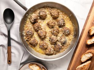 French Onion Meatballs in a Pan at a Table Setting With Toasted Baguette Slices on a Wooden Platter and a Spoon on the Counter, All on a White Table Cloth