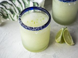 Frozen margarita in a blue rimmed glass with limes in front and another glass and linen behind it.