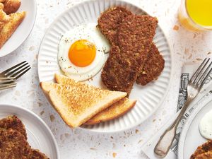 Plate With Sliced Goetta, a Sunny Side Egg, and Toast, and in the Surroundings, a Glass of Orange Juice, Another Plate With a Serving of Breakfast, a Plate With Sliced Goetta, a Plate of Toast, and Forks on the Counter