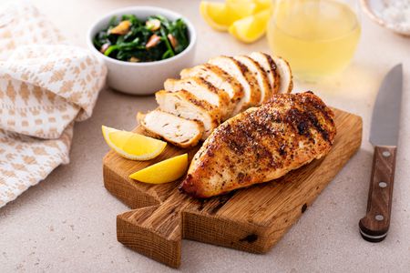 Grilled chicken breasts (one cut into slices, the other whole) on a wooden board with lemon slices, and in the surroundings, a glass of wine, more lemon slices, and a bowl of spinach. 
