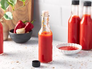 Bottle of Homemade Hot Sauce Open With the Cap Next to the Bottle on the Counter, and in the Surroundings, a Small Bowl of Hot Sauce, More Bottles of Homemade Hot Sauce, a Bowl of Red Fresno and Fresh, Whole Garlic Cloves, and a Kitchen Plant