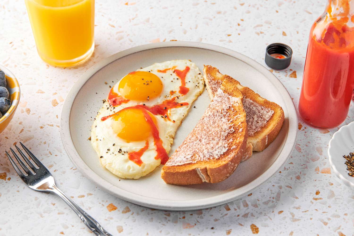 Plate With Two Sunny Side Eggs Topped With Homemade Hot Sauce and Cracked Pepper and Buttered Toast. In the Surroundings, an Open Bottle of Homemade Hot Sauce, a Glass of Orange Juice, a Bowl of Blueberries, and a Fork on the Counter