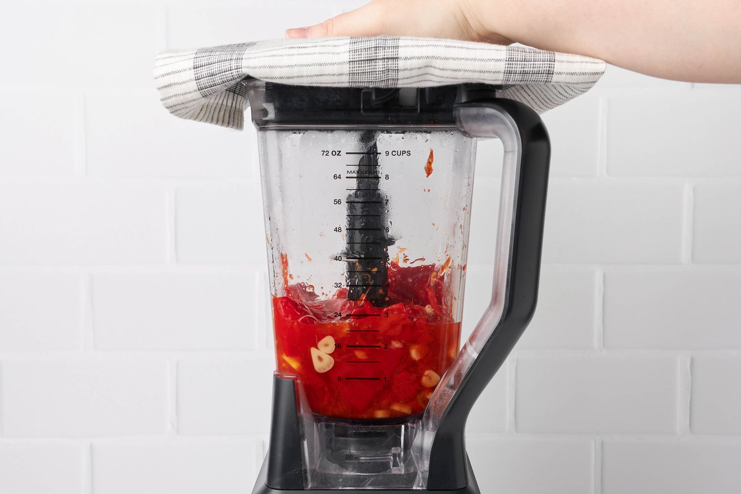 Hot Sauce Mixture in a Blender, and a Hand Holding a Kitchen Towel Onto the Top of the Blender