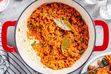 Jollof Rice in a Dutch Oven, and Next to It, a Plate With a Serving of Rice, a Small Saucer, and Glasses of Water