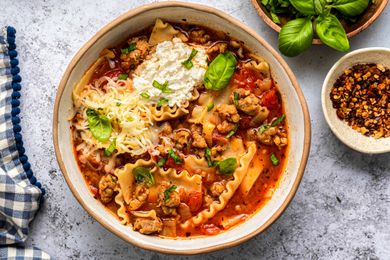 Lasagna Soup in a Bowl With Noodles, Sausage, Ricotta, and Fresh Basil, and in the Surroundings, Another Bowl With Crushed Red Peppers, a Saucer With Basil Leaves, and a Blue and White Checkered Kitchen Towel 