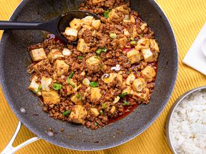 Mapo Tofu in a Pan With a Serving Spoon Next to a Bowl of Rice and a Soup Spoon on a Napkin, All on a Yellow Kitchen Towel