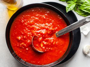 Bowl of Homemade Marinara Sauce With a Ladle, and in the Surroundings, a Bottle of Olive Oil, Garlic Cloves, and Fresh Basil Sprigs