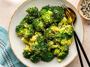 Bowl of Microwave Broccoli with Sesame Dressing with Utensils, Next to a Table Napkin, a Glass of Water, and a Bowl with More Sesame Seeds