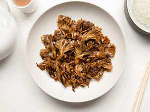 Stir-Fried Maitake Mushrooms with Garlic and Chile Oil in a Plate Next to a Pair of Chopsticks, a Bowl of Rice, a Cup of Tea, and a Tea Kettle