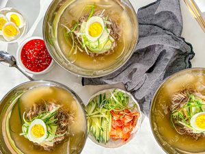 Three Bowls of Mul Naengmyeon Surrounded By Bowls of Toppings (One Bowl With Sliced Cucumbers, Kimchi, and Julienned Green Onions, Another With Spicy Gochujang Sauce, and a Bowl of Halved Hard Boiled Eggs), a Grey Kitchen Towel, and Utensils (a Spoon and Pair of Chopsticks)