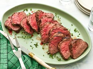 Sliced Oven Roasted Fillet of Beef on a Plate, and in the Surroundings, the Carving Utensils on the Green Kitchen Towel, Two Glasses of Water, and a Stack of Plates