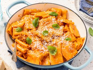 Pot of Paccheri With Quick Sausage Ragu With a Spoon, and in the Surroundings, a Glass, a Block of Parmesan, a Stack of Plates, and Basil Leaves