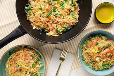Pancit Bihon (Filipino Rice Noodles) in a Wok and Two Servings in Two Bowls at a Table Setting with Utensils and an Empty Bowl