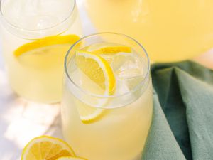Two Glasses of Lemonade With Ice and Lemon Slices Next to a Pitcher With More Lemonade, Halved Lemons on the Counter, and a Sage Table Napkin 