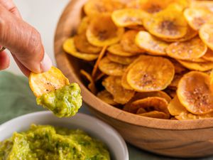 Hand Dipping a Plantain Chip in a Bowl of Guacamole, and in the Background, a Large Bowl Filled With Plantain Chips