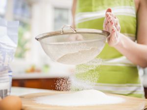 a woman sifting flour onto a counter using a mesh strainer
