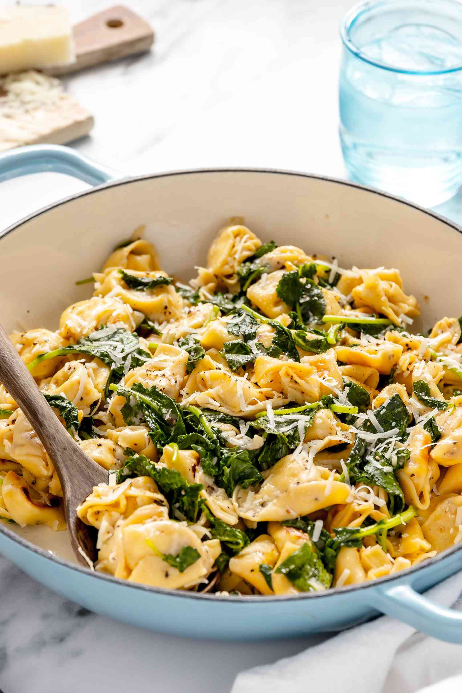 Skillet Cacio e Pepe Tortellini With Wilted Greens in a Pot With a Serving Spoon, All on a White Kitchen Towel on the Counter, and in the Background, a Glass of Water and a Block of Pecorino Romani Cheese on a Cheese Board 