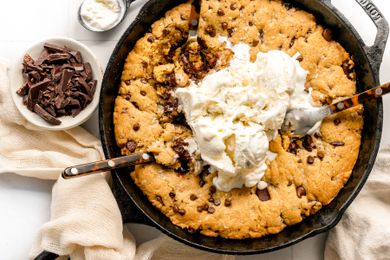 Skillet Chocolate Chip Cookie Topped with Ice Cream with Spoons Digging into the Dessert, Surrounded by a Bowl of Chocolate and an Ice Cream Scoop with More Ice Cream