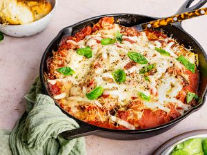 Skillet Eggplant Parmesan Topped With Fresh Basil in a Cast Iron Skillet With a Spoon, and in the Surroundings, a Bowl of Salad Greens, a Bowl of Garlic Bread, and a Mint Kitchen Linen on the Handle of the Cast Iron Skillet